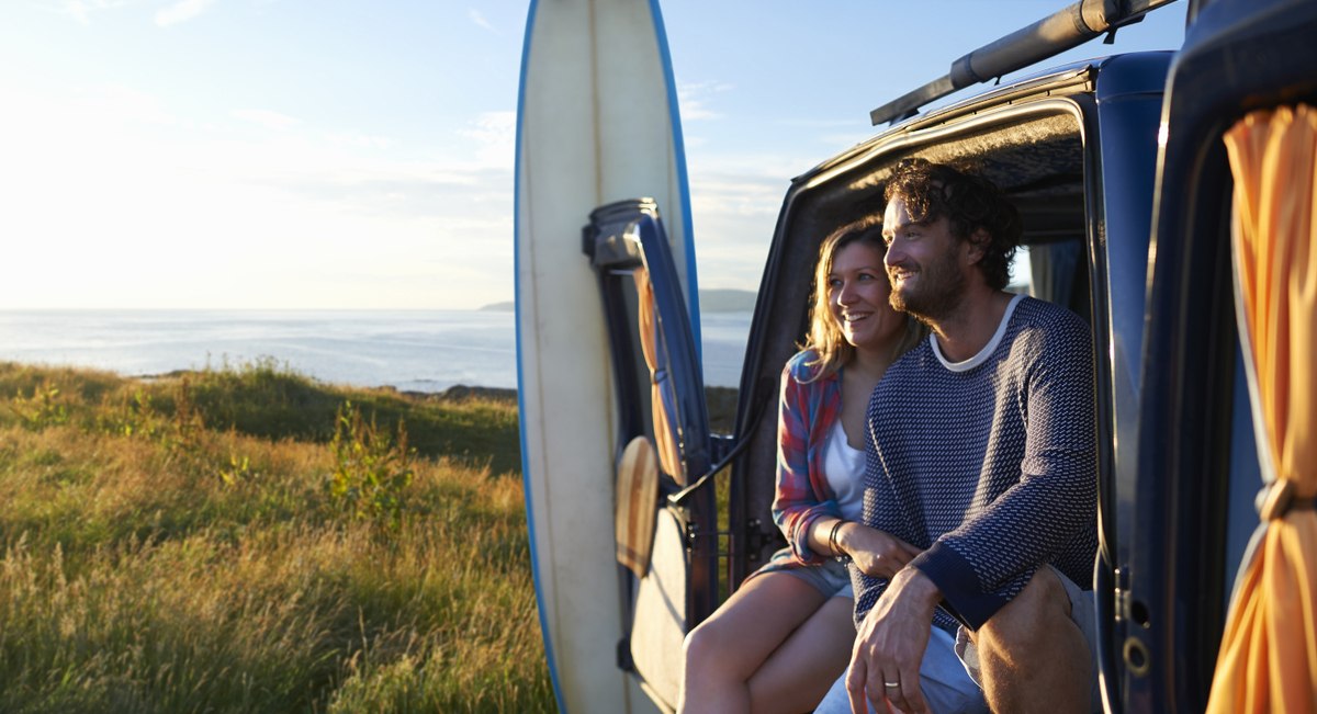 Ein junges Paar, Mann und Frau, sitzen in der offenen Schiebetür ihres Campingbusses auf einer Wiese