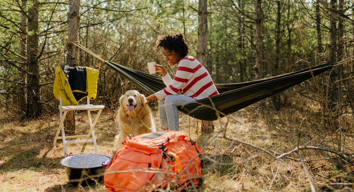 Eine Frau sitzt in einer Hängematte beim Camping im Wald mit einem Hund.